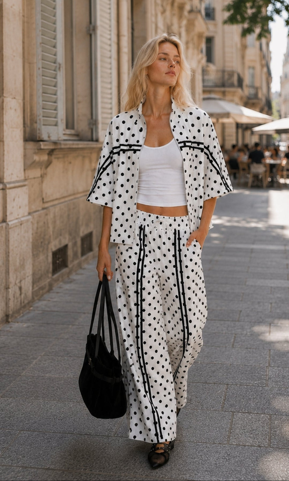 Blonde Frau läuft entspannt durch die Stadt und trägt ein weißes Polka-Dots-Set mit schwarzer Schultertasche.
