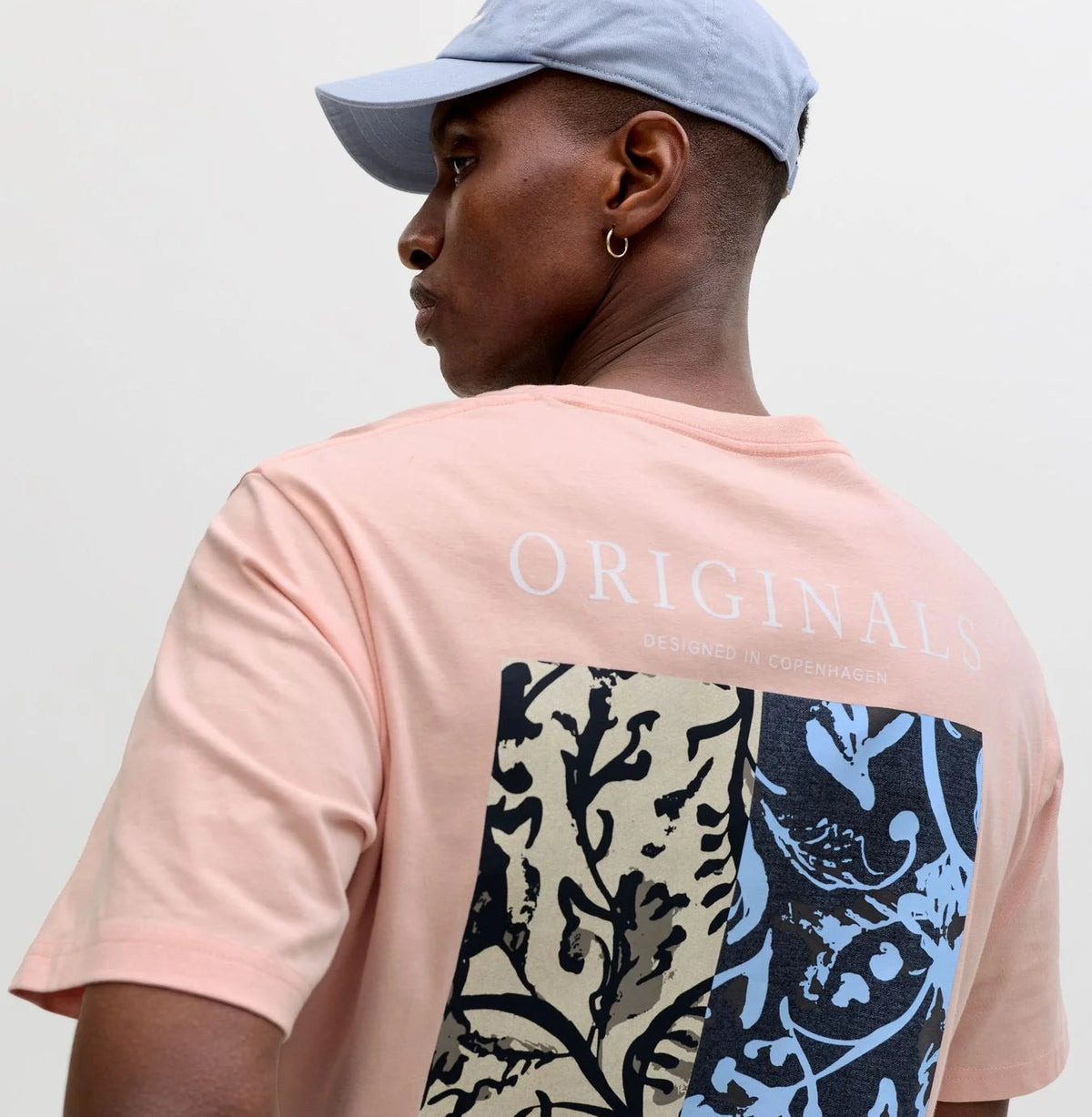 Rosa Herren-T-Shirt mit großem Print auf der Rückseite, getragen mit hellblauer Cap vor hellem Studiohintergrund.