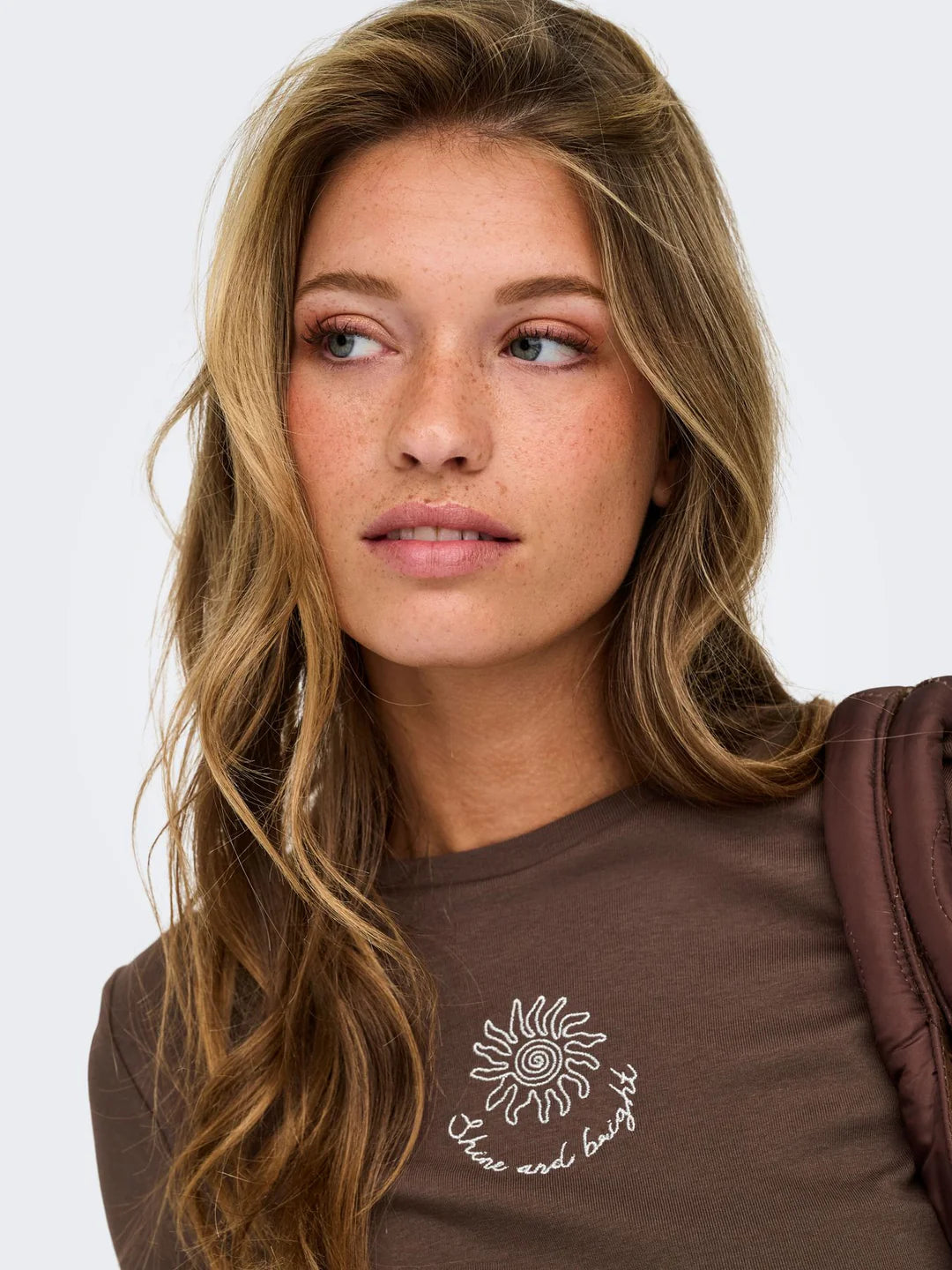 Braunes Damen-T-Shirt mit kleiner Stickerei auf der Vorderseite, kombiniert mit einer braunen Schultertasche vor hellem Studiohintergrund.